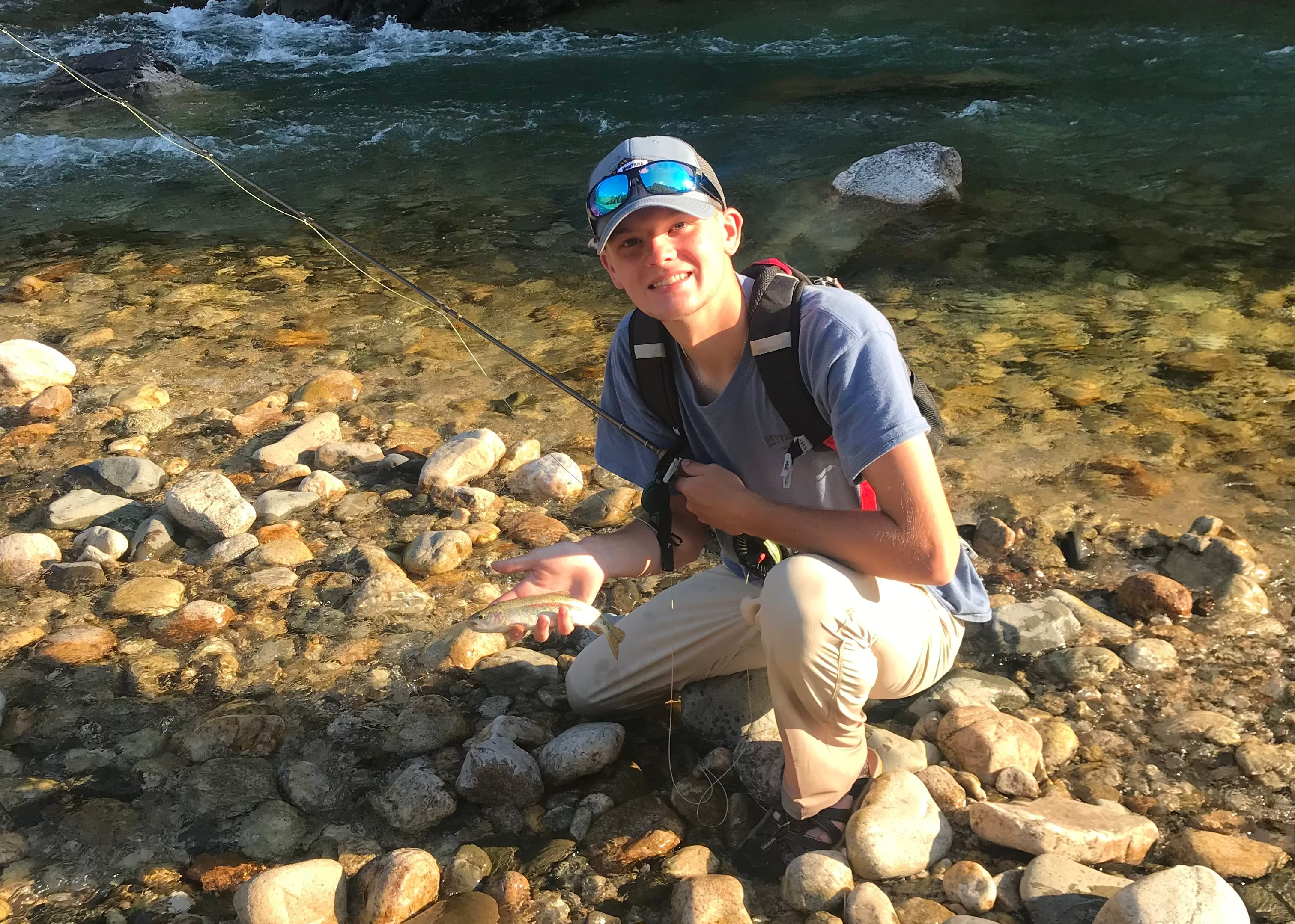 Person sitting on rocks by a river holding a fish, wearing a cap and sunglasses.
